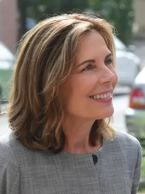 Portrait of a person with shoulder-length wavy hair smiling in profile, wearing a light gray top, photographed outdoors with soft green foliage blurred in the background.