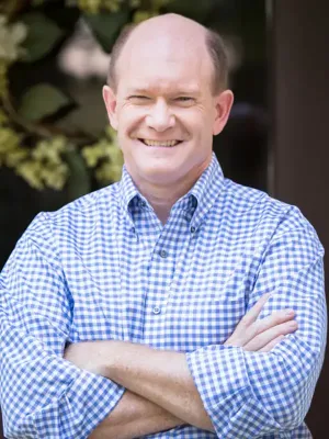 Professional headshot of a man smiling with arms crossed, wearing a blue and white checkered shirt, with a soft-focus green background.