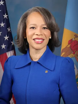 Official portrait of a woman wearing a blue blazer with a gold badge, smiling in front of the U.S. flag and an organizational flag backdrop.